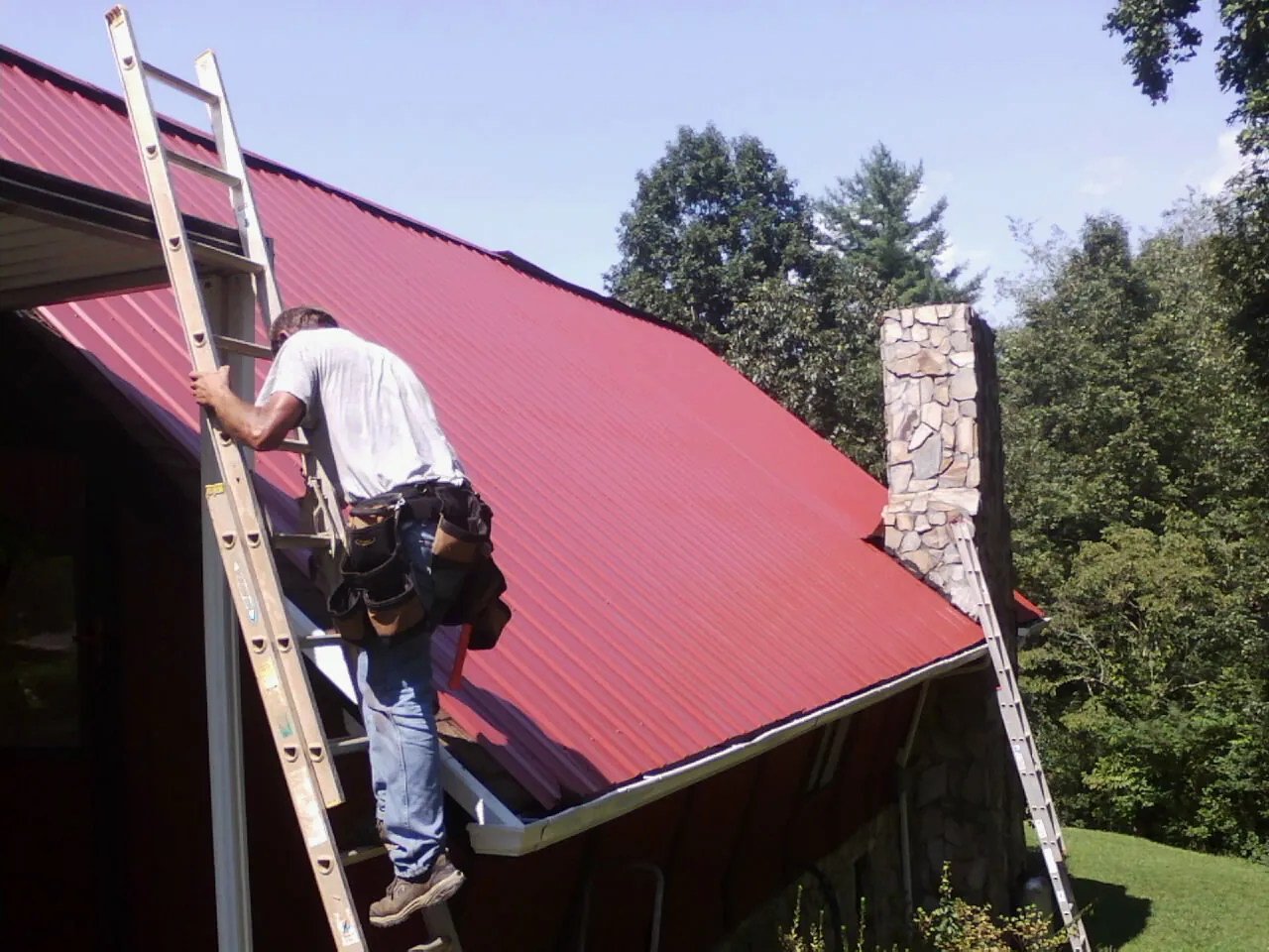 Roofer on a ladder beside a red metal roof and stone chimney — Soffit & Fascia Repair by Roofing Brothers in Turtle Creek